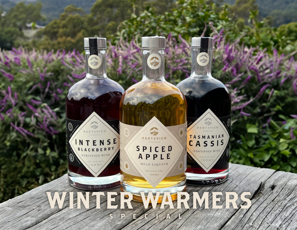 3x700ml bottles of Hartzview fortified wines sitting on a wooden bench with purple flowers in the background. Overlaid over the picture is the text Winter Warmers Special.